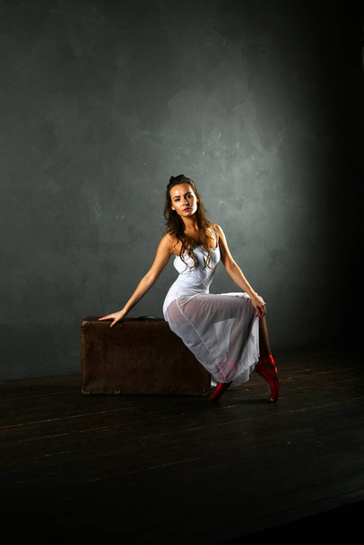ballerina in een witte jurk en rode punten zit op een koffer op een grijze achtergrond - Foto, afbeelding