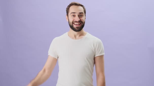 A friendly young man wearing white t-shirt is waving his hand doing hello gesture while standing isolated over gray background - Video