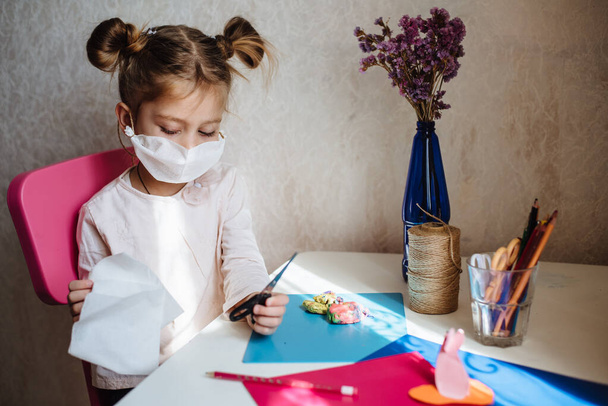 Pięcioletnia dziewczynka w masce medycznej zajmuje się twórczą pracą. Maska ochronna własnymi rękami. Co zrobić, aby poddać dziecko kwarantannie. Nauka w domu podczas kwarantanny. Zostań w domu.. - Zdjęcie, obraz