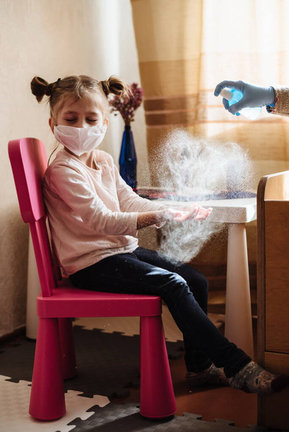 Mom sprays a disinfectant antibacterial spray on the girl's hands. Preventive measures against Covid-19 infection. Sick child in protective mask on isolation at home. - Photo, Image