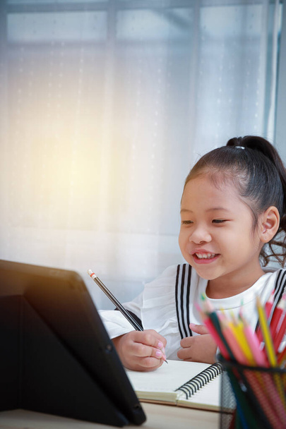 Aasialainen tyttö on onnellinen kotitehtäviä verkossa kotona. Etäopetus lapsille Coronaviruksen aikana. Hän hymyilee onnellisena kirjoittaessaan kirjaan ja kommunikoidessaan opettajan kanssa videopuhelulla
. - Valokuva, kuva