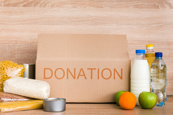 Charity donation box made of cardboard with food with oil, milk and water, herbs, apples and oranges, cereals and canned food, rice and pasta. Beige wooden background - Photo, Image