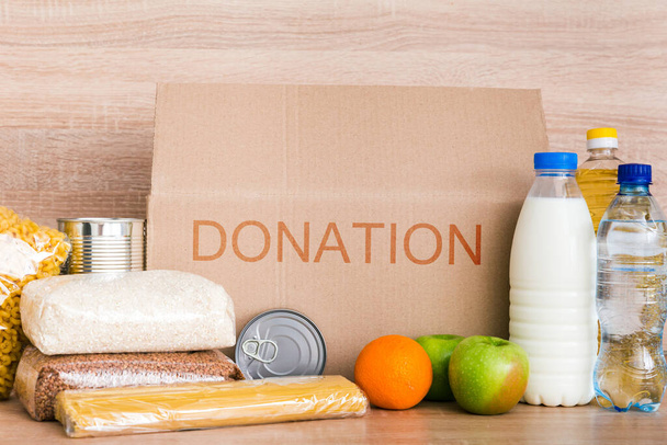 Caixa de doação de caridade feita de papelão com alimentos com óleo, leite e água, ervas, maçãs e laranjas, cereais e alimentos enlatados, arroz e massas. Fundo de madeira bege
 - Foto, Imagem