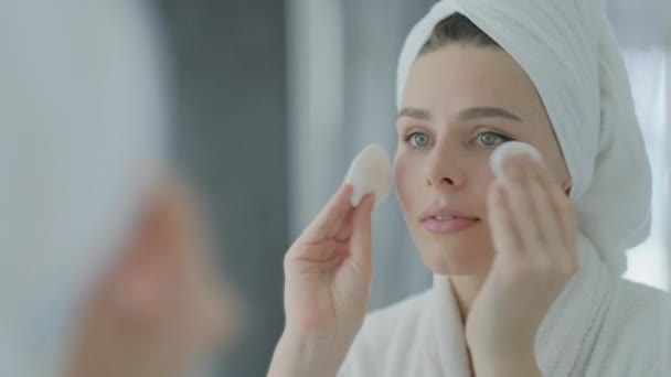Beautiful woman in white towel on her head and a bathrobe looking in reflection in the mirror and cleansing face skin removing make up with cotton disk, skincare concept. - Materiał filmowy, wideo