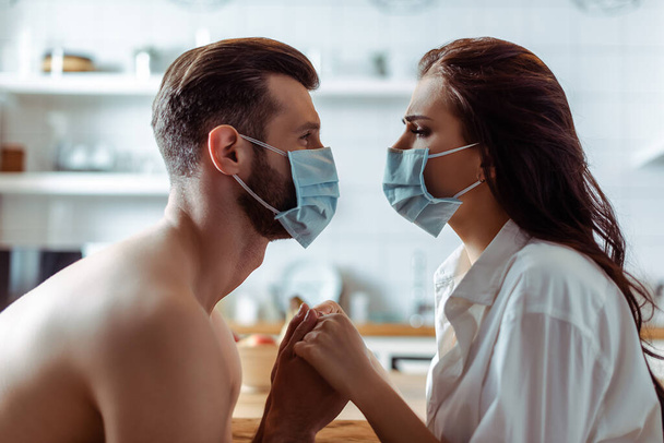 side view of young couple in medical masks holding hands in kitchen - Foto, afbeelding