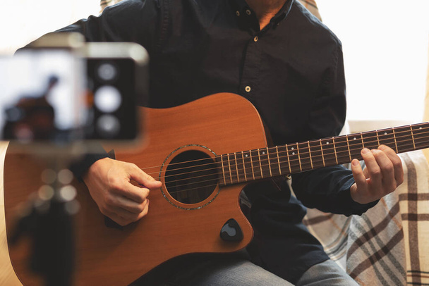 Man with acoustic guitar in a shirt and cap plays live staying home for the audience in front of the smartphone camera during quarantine and forced self-isolation due to coronavirus - Photo, image
