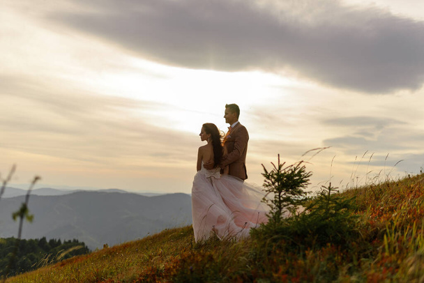 Braut und Bräutigam umarmen sich zärtlich. Sonnenuntergang. Hochzeitsfoto vor dem Hintergrund herbstlicher Berge. Starker Wind bläst Haare und Kleid auf.  - Foto, Bild