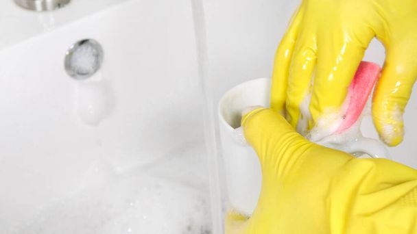 Person in yellow rubber gloves washer a mug in kitchen sink - Foto, afbeelding