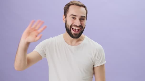 A friendly positive young man wearing a white t-shirt is waving his hand doing hello gesture with a smile standing isolated over gray background - Felvétel, videó