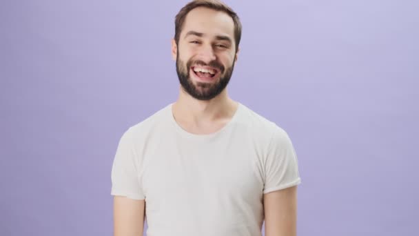 A smiling emotional young man wearing a white t-shirt is showing a thumb up gesture to the camera standing isolated over gray background - Кадры, видео