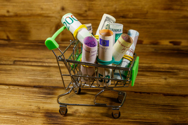 Shopping cart with money from different countries: euros, us dollars, ukrainian hryvnia, russian rubles, egyptian pounds on wooden background - Foto, immagini