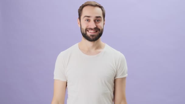 A happy playful young man wearing a white t-shirt is pointing down standing isolated over gray background - Video