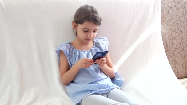 Curious cute girl preschool child using a digital technology device looks telephone. Plays in the phone sitting on the sofa alone. Children sataying at home during quarantine COVID-19 - Кадри, відео
