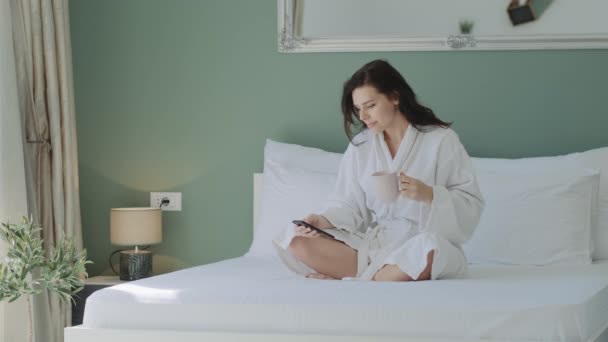 Brunette woman in a white bathrobe looking at phone, drinking tea or coffee while sitting on bed. Pretty girl holding mug and reading digital news at home. - Felvétel, videó