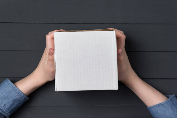 Children's hands holds white square cardboard box. Black background. Top view. Shipping concept - Fotó, kép