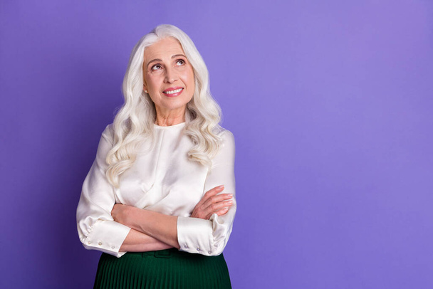 Photo of attractive aged white haired lady smiling good mood arms crossed look up empty space dreamy wear white blouse green long midi skirt isolated pastel purple color background - Foto, afbeelding