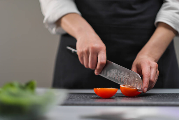 Een vrouwelijke kok in een wit uniform en een zwart schort in de keuken van het restaurant. De kok snijdt de groenten met een groot keukenmes. - Foto, afbeelding