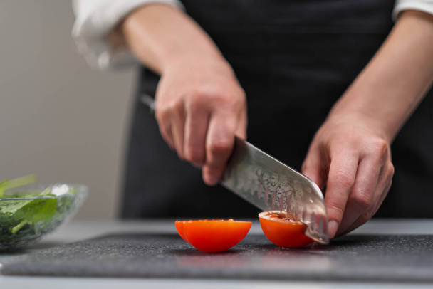 A female chef in a white uniform and a black apron in the restaurant kitchen. The cook cuts the vegetables with a large kitchen knife. - Foto, Bild