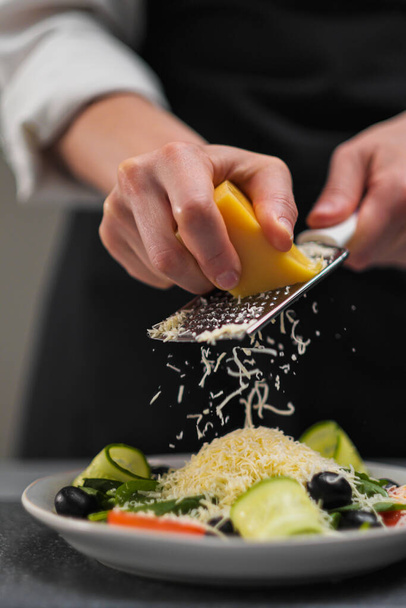 A female chef in a white uniform and a black apron in the restaurant kitchen. Cooking a salad. The cook rubs the parmesan cheese on a small grater. - Фото, зображення