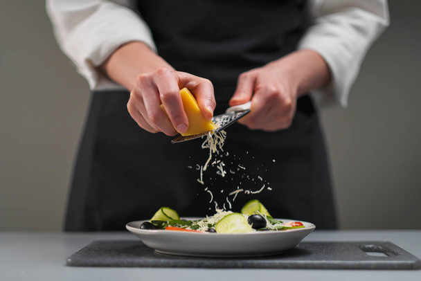 A female chef in a white uniform and a black apron in the restaurant kitchen. Cooking a salad. The cook rubs the parmesan cheese on a small grater. - Zdjęcie, obraz
