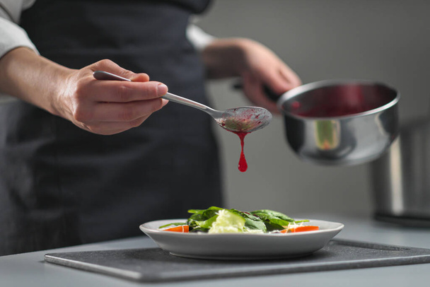 A female chef in a white uniform and a black apron in the restaurant kitchen. The cook pours red cranberry salad sauce. Making and decorating food. - Φωτογραφία, εικόνα