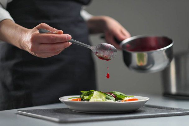 A female chef in a white uniform and a black apron in the restaurant kitchen. The cook pours red cranberry salad sauce. Making and decorating food. - Foto, Imagem
