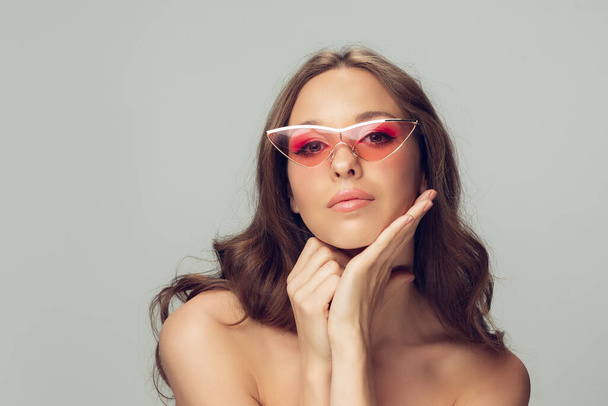 Close up of beautiful young woman with long healthy curly hair and bright make up wearing stylish pink eyewear isolated on grey studio backgroud - Photo, image