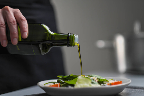 白い制服を着た女性シェフとレストランのキッチンで黒いエプロン。料理。コックは緑のボトルからオリーブオイルを注ぎます. - 写真・画像