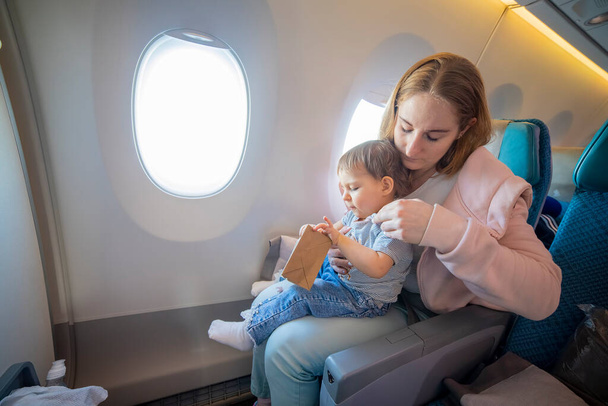 Концепція догляду за мамою. молода красива мати сидить у кріслі літака і вирівнює одяг маленького милого малюка, що сидить на колінах
 - Фото, зображення