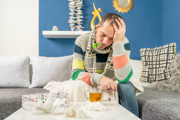 Человек с гриппом и термометром сидит на диване
 - Фото, изображение