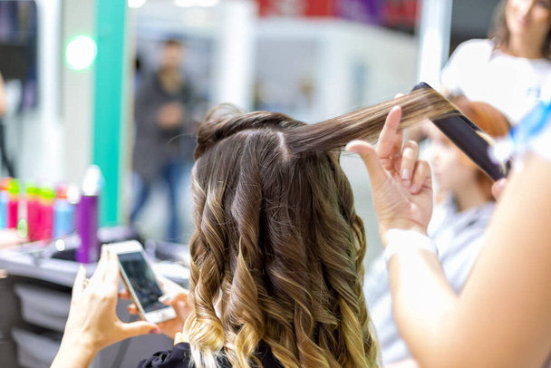 close up stylista dělá dívka klient styling vlasy mává s curling žehličkami v kadeřnictví. zákaznická dívka se dívá do smartphonu v rozmazání. zblízka, měkké zaměření - Fotografie, Obrázek