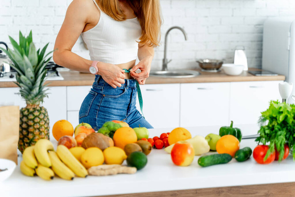 Dieting concept. Beautiful young woman in the kitchen with healthy food. Measuring tape on waist. Fruits and Vegetables - Fotó, kép