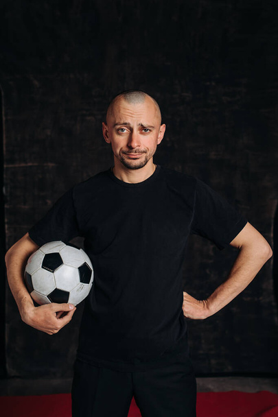 A male sports teacher stands against a dark background, holding a soccer ball, looking at the camera and smiling.portrait of a football fan . A man in a black t-shirt - Fotó, kép
