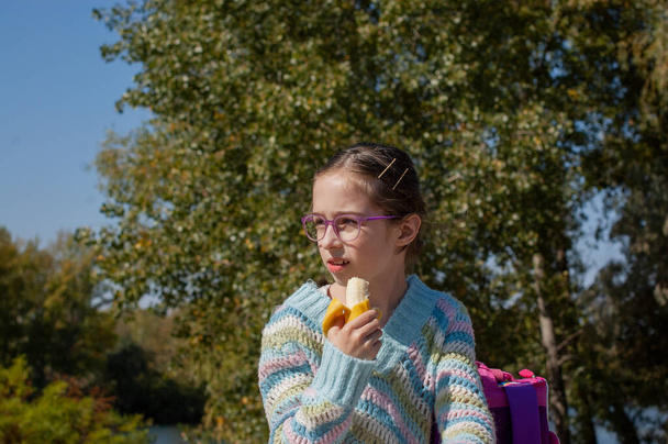 Μια μαθήτρια τρώει μια μπανάνα στο δρόμο. Τα παιδιά της γενιάς Άλφα θα είναι η κινητήρια δύναμη της προόδου του αιώνα μας. Είναι πιο ισορροπημένες, θετικές, λιγότερο επιθετικές.. - Φωτογραφία, εικόνα