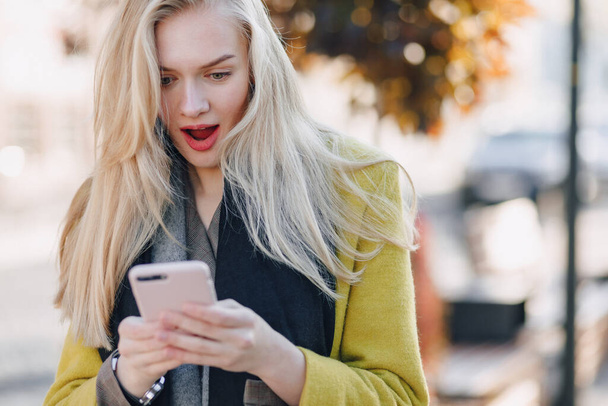 Nette emotionale attraktive blonde Frau im Mantel mit Smartphone geht durch die Straßen der Stadt. Kommunikation während des Spaziergangs, Lebensstil, Straße bei gutem Wetter. - Foto, Bild