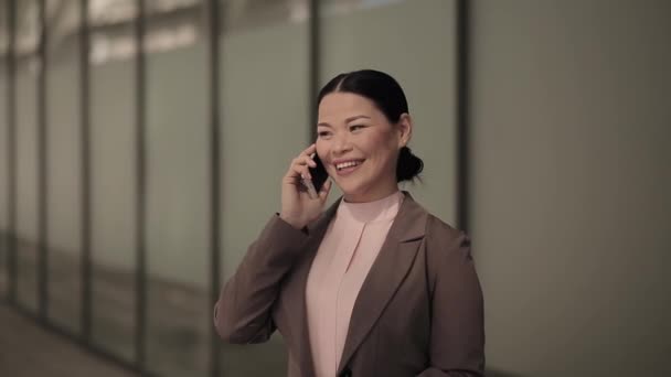 Asian businesswoman talking on the phone wearing business suit stand outdoors with a business office center on background. Business concept. Prores 422 - Кадры, видео