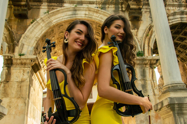 Dvě krásné dvojčata houslistky ve žlutých koncertních šatech pózují s elektrickými houslemi v blízkosti Hadrianovy brány ve starém městě Antalya Kaleici Turkey. Stock image. - Fotografie, Obrázek