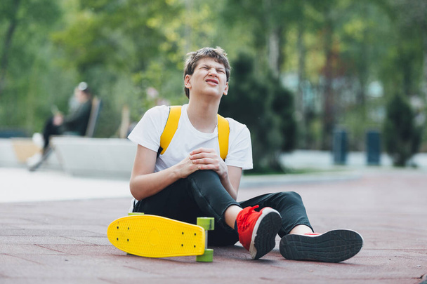 Teenager im roten T-Shirt trainieren in ihrer Freizeit im Stadtpark Schlittschuhlaufen. Junge stürzte vom Schlittschuh und verletzte sich beim Tricksen am Knie. Kind im Freien mit Skateboard. Hobby, Sport für Kinder. - Foto, Bild