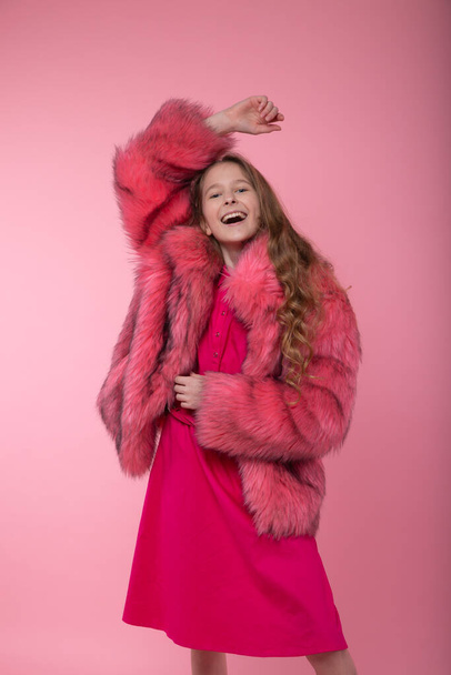 Cheerful girl model in fashionable clothes, a light pink fur coat, pink bright dress and white sneakers posing holding her hand up on a pink background. - 写真・画像