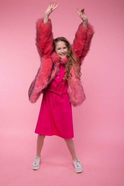 A beautiful young girl dances and raises her hands up in a pink eco-coat and white sneakers on a pink background. Clothing style. - Foto, immagini