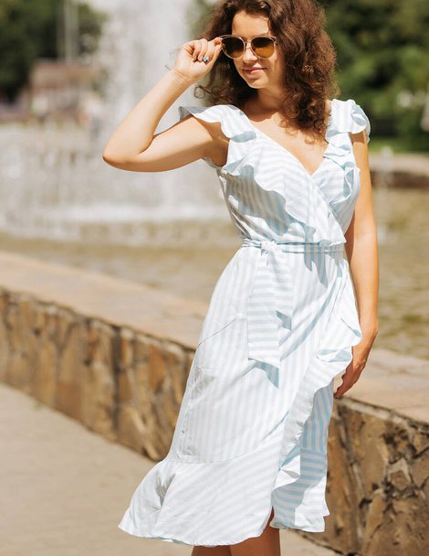 Dziewczyna w okularach przeciwsłonecznych w pobliżu fontanny na nabrzeżu w niebieskiej sukience - Zdjęcie, obraz