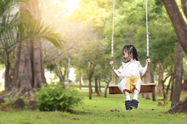 Маленька дівчинка гойдається на дитячому майданчику в літньому саду в теплий і сонячний день на відкритому повітрі
. - Фото, зображення