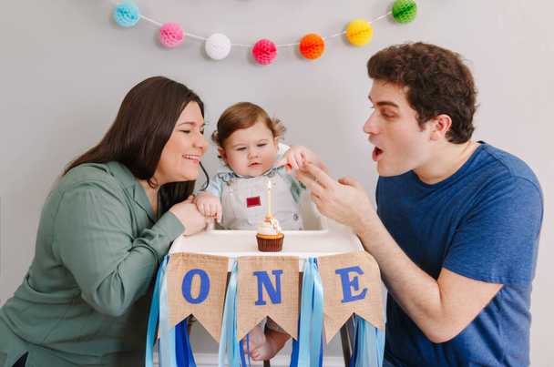 Blanke familie met kleine jongen die zijn eerste verjaardag thuis viert. Moeder vader vader met kind peuter blaast kaars op cupcake. Gelukkige verjaardag en taart smash concept. - Foto, afbeelding