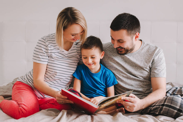 Rodzina trojga siedząca na łóżku w książce do czytania. Matka, ojciec i syn spędzają razem czas w domu. Rodzice rozmawiają z dzieckiem. Dzień rodzinny autentyczny styl życia. - Zdjęcie, obraz