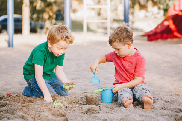 Twee blanke kinderen zitten in een zandbak te spelen met strandspeelgoed. Vriendjes die plezier hebben op de speelplaats. Zomer buitenactiviteiten voor kinderen. Vrije tijd levensstijl kindertijd. - Foto, afbeelding