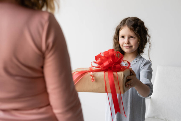 Birthday concept. Girl schoolgirl in a blue dress gives her mom a gift. Her mom has a birthday. She is smart and happy. The box with a surprise is big and has a red bow on it. - Fotografie, Obrázek