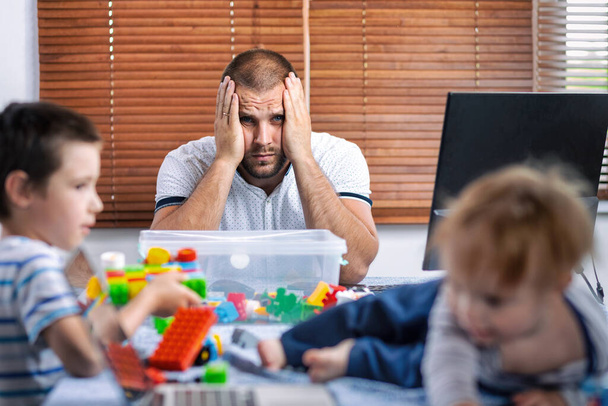 Dad with two little boys on his knees tries to laugh at home . A young woman looks after children and works on a computer. Father does home business, soft focus - Photo, Image