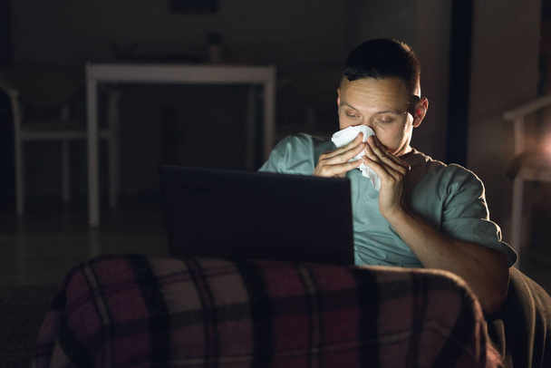 Ένας άρρωστος άντρας κάθεται μόνος στο σπίτι του στην απομόνωση. Να δούμε ταινία το βράδυ. Τρέχει μύτη και θερμοκρασία σε έναν άντρα. Φύσα τη μύτη σου σε χαρτοπετσέτες. Θέση για κείμενο - Φωτογραφία, εικόνα