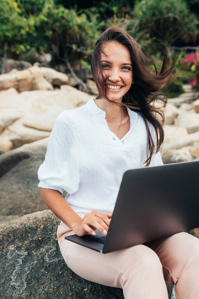 Young successful, smiling girl working using a laptop, sitting on a rocky seashore. Business woman working on remote work while traveling, blogger, freelancer, compiles content plan, work online. copy space - Photo, Image