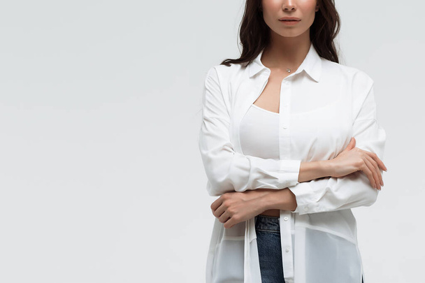 Crop zelfverzekerde langharige vrouw in wit shirt en lingerie en jeans met gekruiste handen op witte achtergrond - Foto, afbeelding